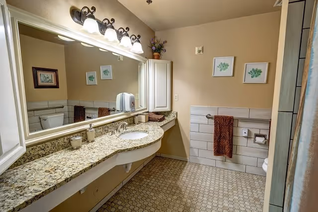 A bathroom with a large granite countertop and an under-mount sink. Above the sink is a large mirror with four light fixtures. The walls are beige with two framed botanical prints. There is a white cabinet mounted on the wall and a towel rack with a brown towel. The floor has small hexagonal tiles, and a toilet is partially visible on the left side.