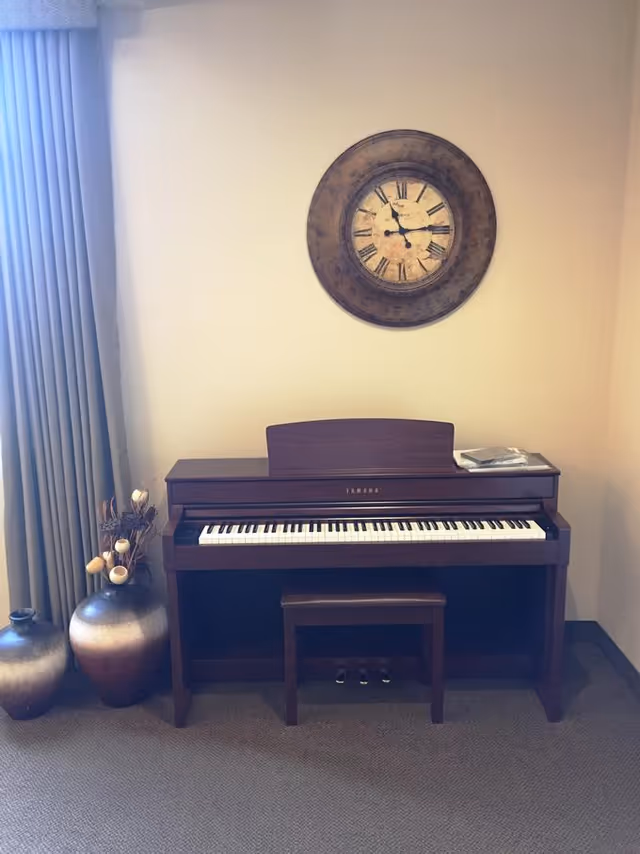 A wooden upright piano with a matching bench placed against a beige wall. Above the piano hangs a large round wall clock with Roman numerals. To the left of the piano are two decorative vases and a curtain covering a window.