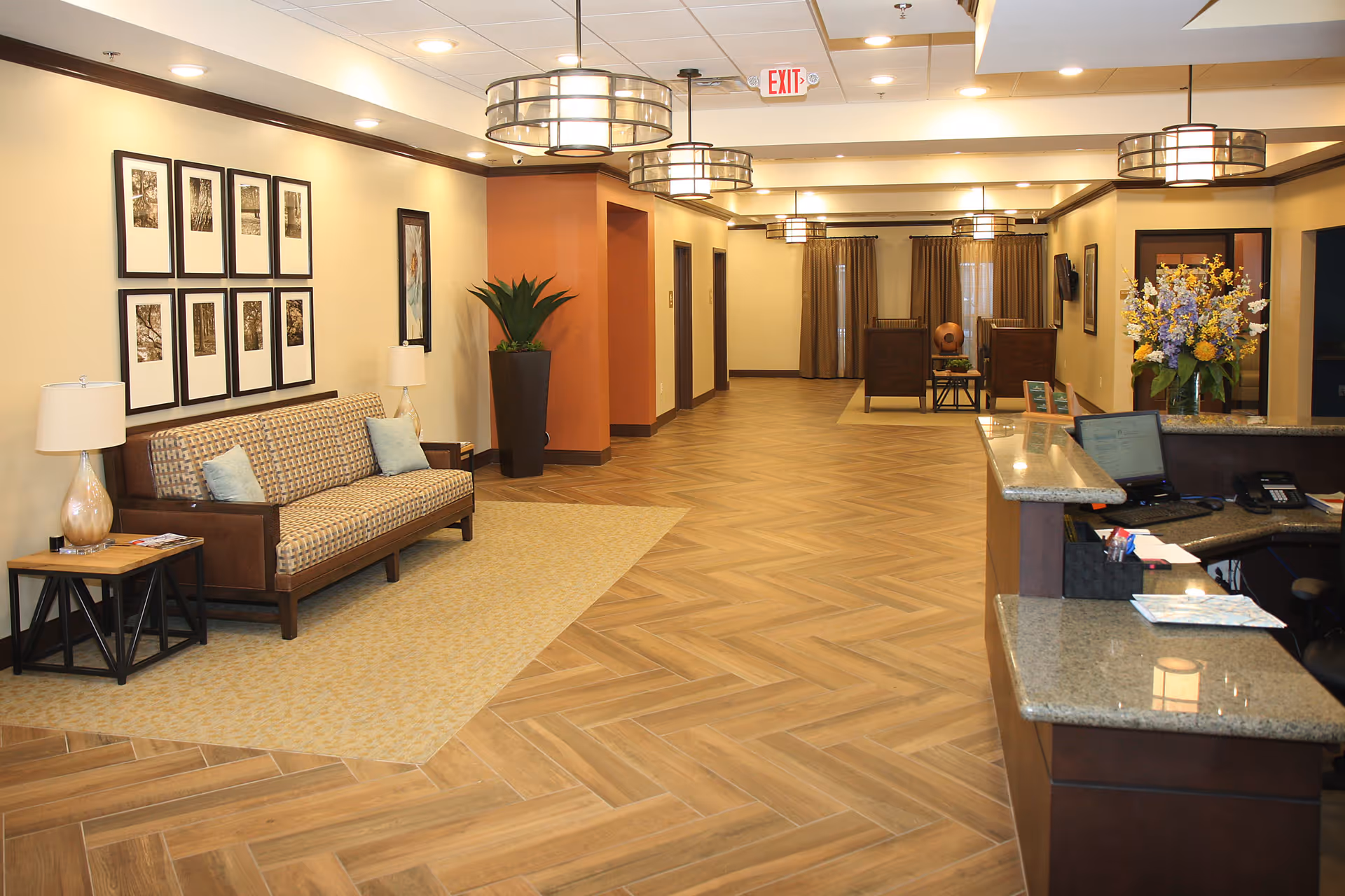 A spacious and well-lit assisted living facility lobby with a patterned wooden floor and beige walls. On the left side, there is a cushioned sofa with two light blue pillows, a side table with a lamp, and framed artwork on the wall above. In the center and background, there are additional seating areas with chairs and a table. On the right side, there is a reception desk with a computer, phone, and a vase of colorful flowers. Ceiling lights and an exit sign are visible overhead.