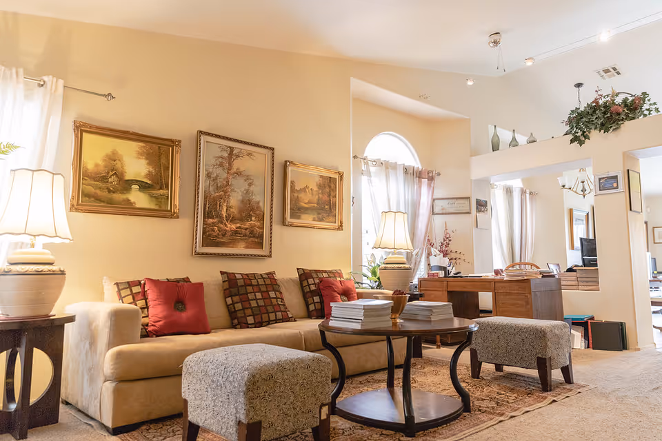 A cozy living room in an assisted living facility with a beige sofa adorned with red and patterned cushions, two upholstered ottomans, a wooden coffee table with stacks of magazines, and two table lamps. The walls feature three framed landscape paintings, and there is a large window with sheer curtains letting in natural light. In the background, a wooden desk with office supplies and a computer is visible, along with decorative plants and vases on a high shelf.