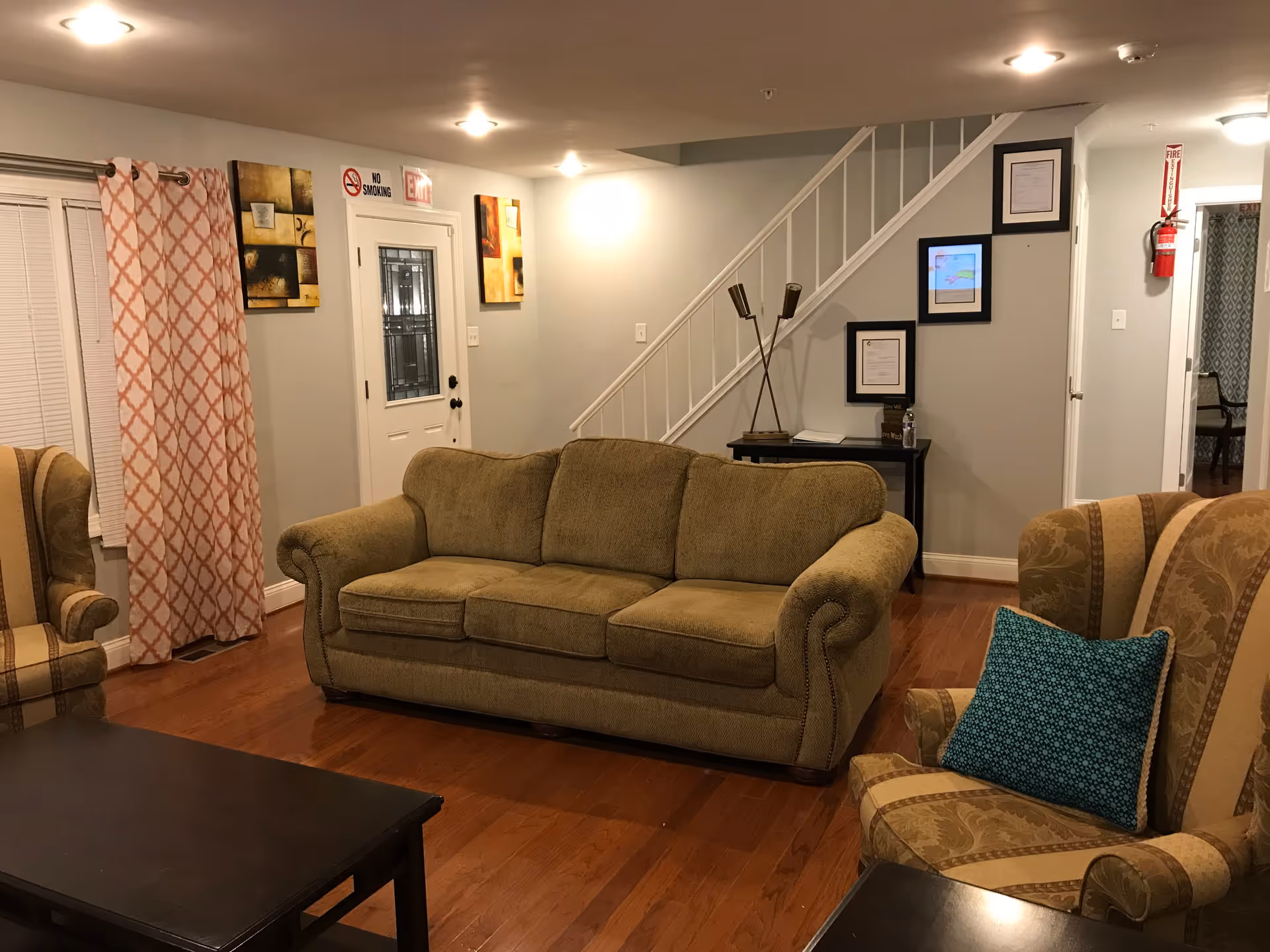 Cozy living room with a green upholstered sofa, patterned armchairs, coffee table, staircase, and front door in a senior living facility.