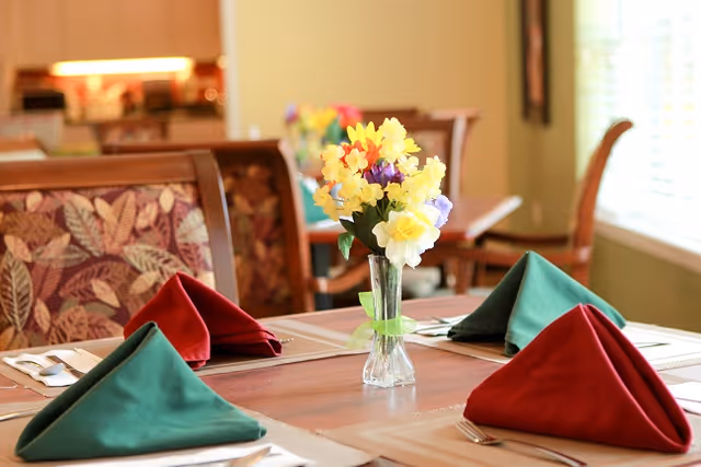 A dining table set with red and green folded napkins, silverware, and a clear vase with yellow, purple, and white flowers in the center. The background shows cushioned chairs with a leaf pattern and a softly lit interior space.