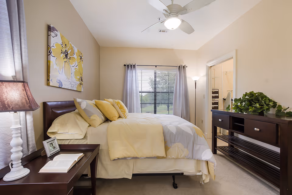 A cozy bedroom with a bed made up with yellow and gray floral bedding, a bedside table with a lamp, an open book, and a framed photo. There is a large window with gray curtains letting in natural light, a ceiling fan above, and a dark wooden dresser with a green plant on top. The room has beige walls and carpeted floor.