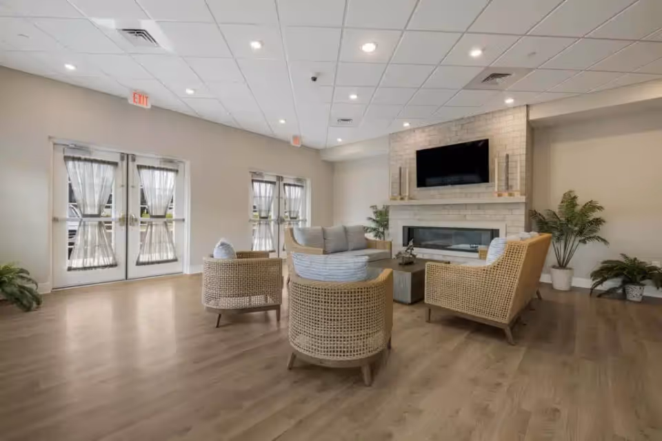 Bright communal living room with wicker seating arranged around a fireplace and wall-mounted TV.