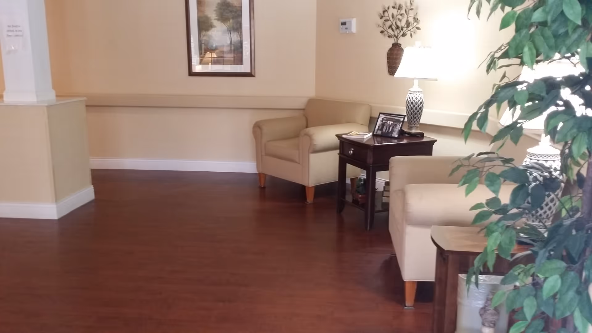A furnished seating area with beige armchairs, side tables, lamps, wall art and a potted plant on wood flooring.