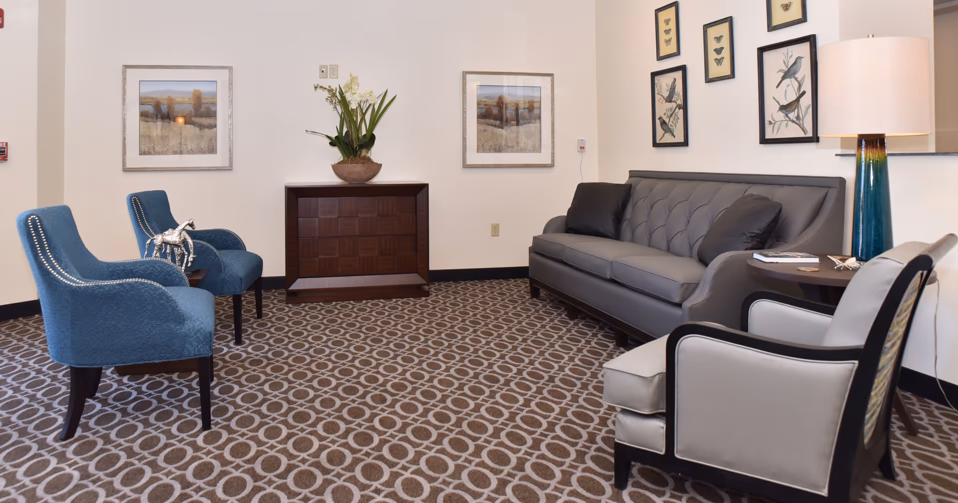 Lobby-style sitting area with blue upholstered chairs, a gray tufted sofa, patterned carpet, framed artwork and a floor lamp.