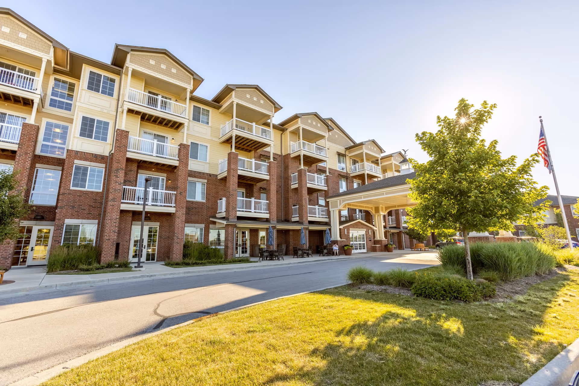 Exterior view of a multi-story senior living facility building with balconies, a covered entrance, landscaping including a tree and grass, and an American flag on a flagpole under a clear sky.