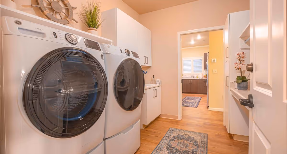 Laundry room with a front-loading washing machine and dryer side by side, white cabinets above and beside them, a small plant on top of the dryer, and a decorative ship wheel on the wall. The room has wooden flooring and a rug, with a doorway leading to a kitchen area in the background.