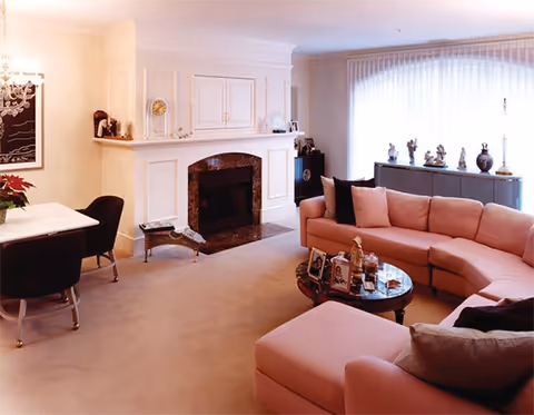 Bright living room with a pink sectional sofa, coffee table, fireplace, dining nook, and a large curtained window.