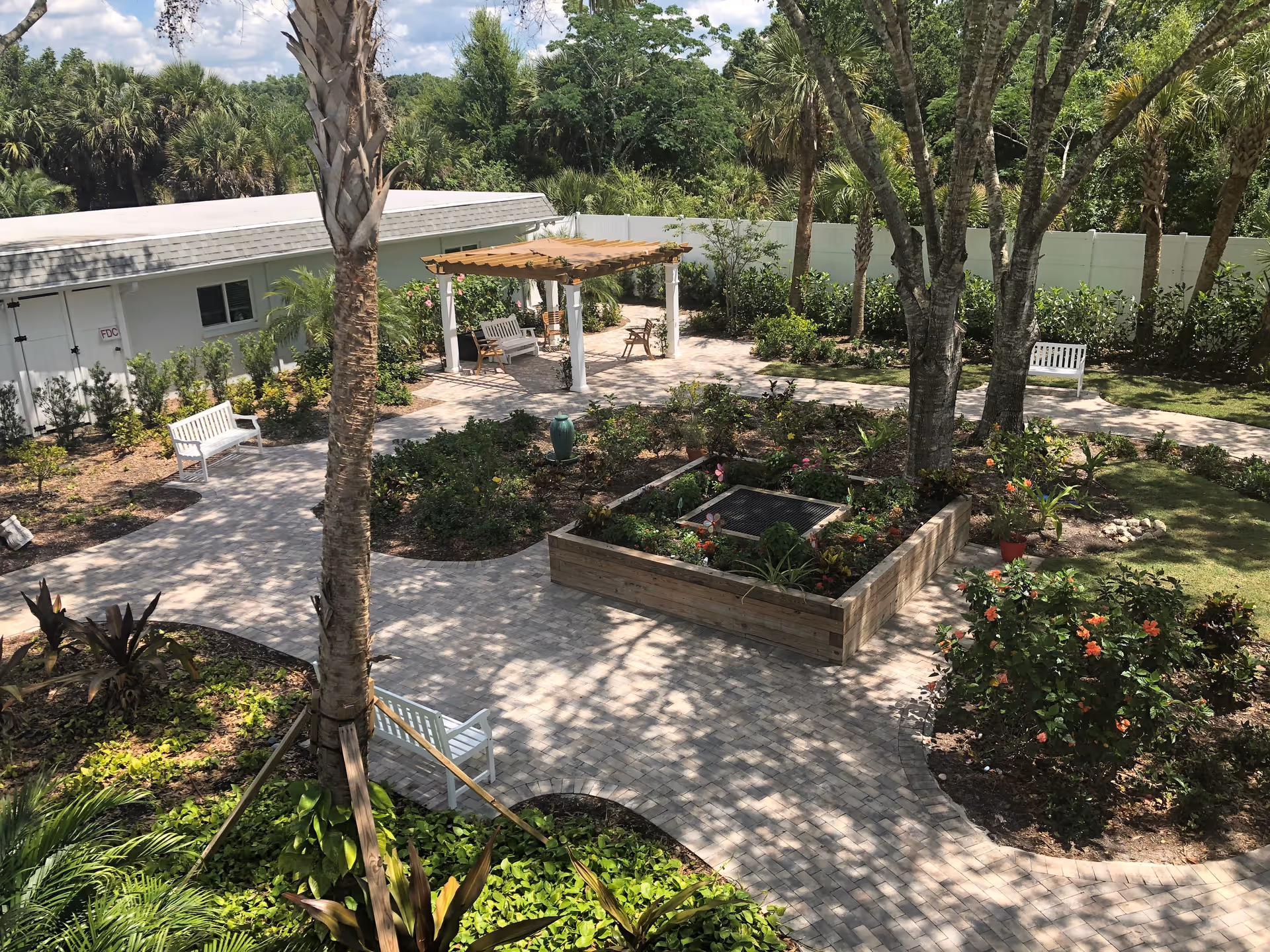 A peaceful outdoor garden area with paved walkways, several white benches, a wooden pergola with seating underneath, various plants and flowers, and tall trees providing shade. A white building and a white fence are visible in the background.