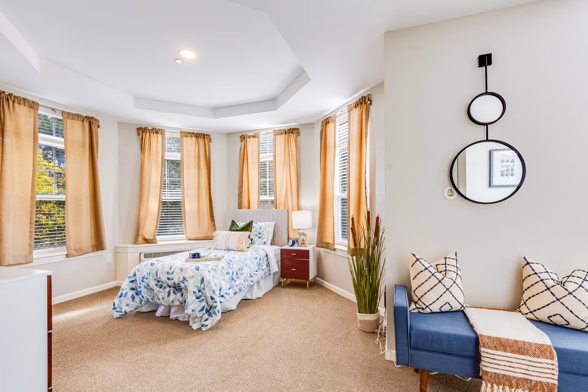 A bright and cozy bedroom with beige walls and carpet. The room features a single bed with a blue and white floral comforter, a green pillow, and a beige bolster pillow. There is a wooden nightstand with a lamp and a framed photo next to the bed. The room has several windows with beige curtains allowing natural light to enter. On the right side, there is a blue cushioned bench with two patterned pillows and a striped throw blanket. Above the bench, two round mirrors are mounted on the wall, one smaller above a larger one. A tall potted plant is placed near the bench.