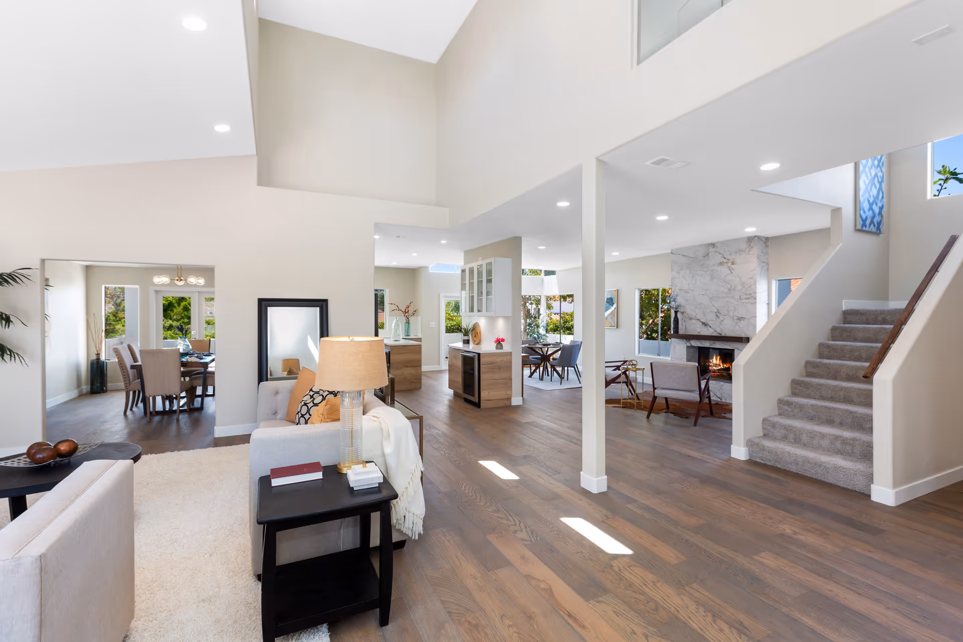 Bright and spacious open-concept living area with light-colored walls and wood flooring. The room features a cozy seating area with a lamp and side table, a fireplace with a marble surround, a dining area with a table and chairs, and a staircase with carpeted steps leading upstairs. Large windows allow natural light to fill the space.