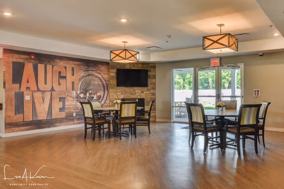 A bright and spacious common area in a senior living facility with wooden flooring, two round tables each surrounded by four chairs with striped upholstery. The wall features a large decorative mural with the words 'LAUGH LIVE' and a rustic design. A flat-screen TV is mounted on a stone accent wall. Large glass doors lead to an outdoor area with greenery visible outside. Two modern pendant lights hang from the ceiling.