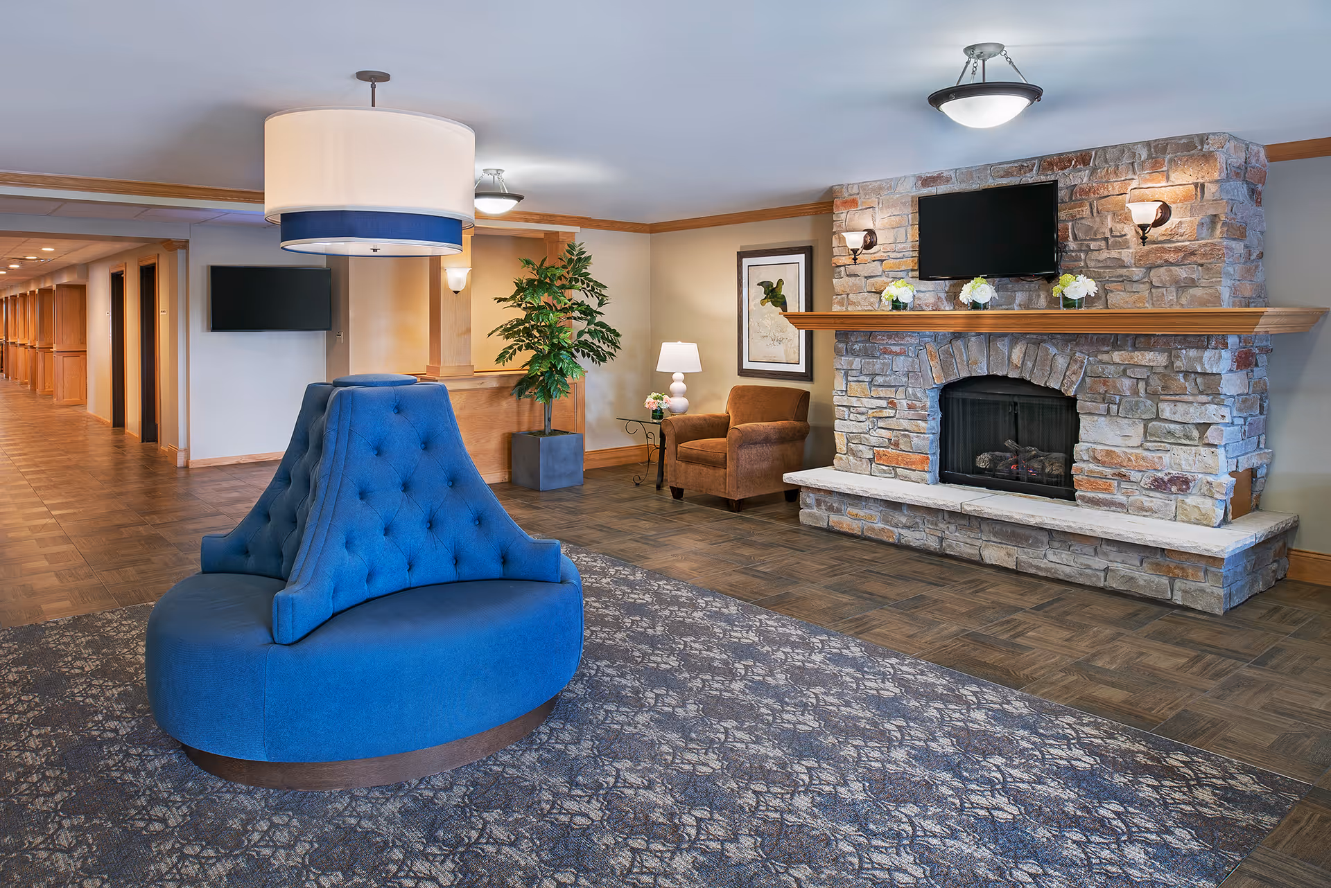 A cozy senior living facility common area featuring a circular blue upholstered seating arrangement in the center, a stone fireplace with a mounted TV above it, a brown armchair next to a small table with a lamp and flowers, a large potted plant, and a hallway leading to rooms on the left side.