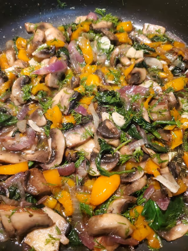 Close-up view of a pan with sautéed vegetables including mushrooms, yellow bell peppers, red onions, spinach, and herbs.