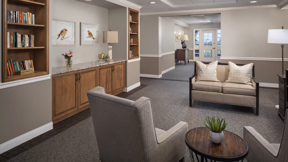 A cozy senior living facility common area with two beige armchairs and a matching loveseat arranged around a small wooden coffee table with a potted plant. The room features built-in wooden bookshelves filled with books, a granite countertop with decorative items, framed bird artwork on the wall, and soft lighting from lamps. The space has neutral-colored walls and carpeted flooring, with a hallway leading to a door with windows.