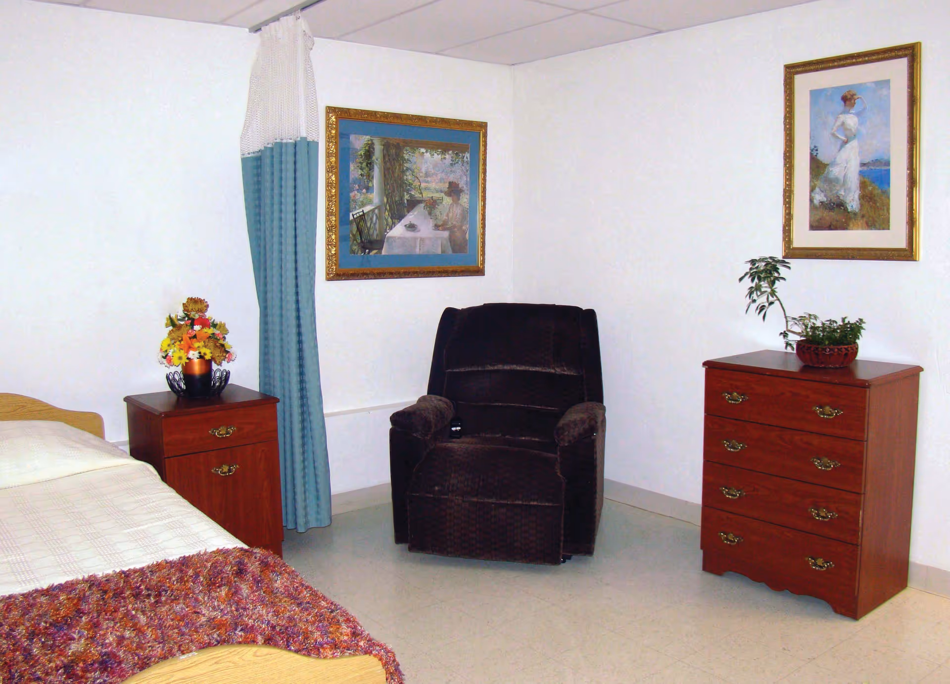 A simple bedroom in a nursing center featuring a single bed with a beige blanket and a colorful throw, a wooden nightstand with a flower arrangement, a dark upholstered armchair, a wooden chest of drawers with a small potted plant on top, and two framed paintings on the white walls.