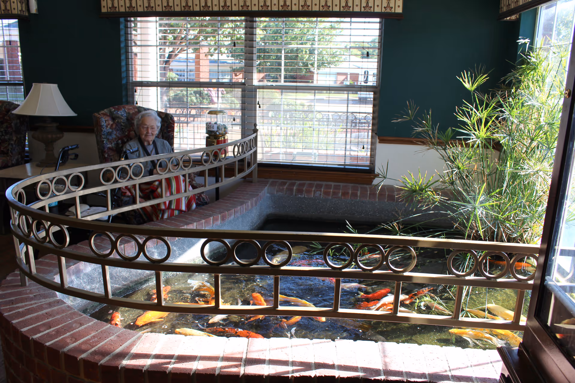 An elderly woman sitting in a floral armchair next to an indoor koi pond surrounded by a decorative metal railing and brick edging. The room has large windows with blinds, a table with a lamp, and some indoor plants near the pond.