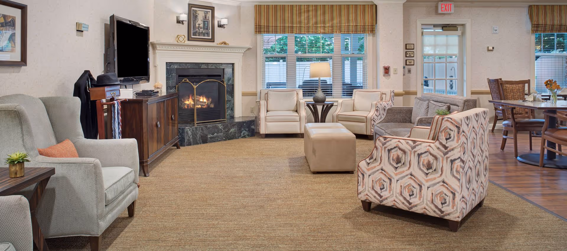A cozy living room area in a senior living facility featuring a lit fireplace with a decorative screen, a flat-screen TV mounted on the wall, various upholstered chairs including patterned and solid colors, a beige ottoman, a side table with a lamp, and large windows with striped valances letting in natural light. In the background, a dining area with wooden chairs and tables is visible.