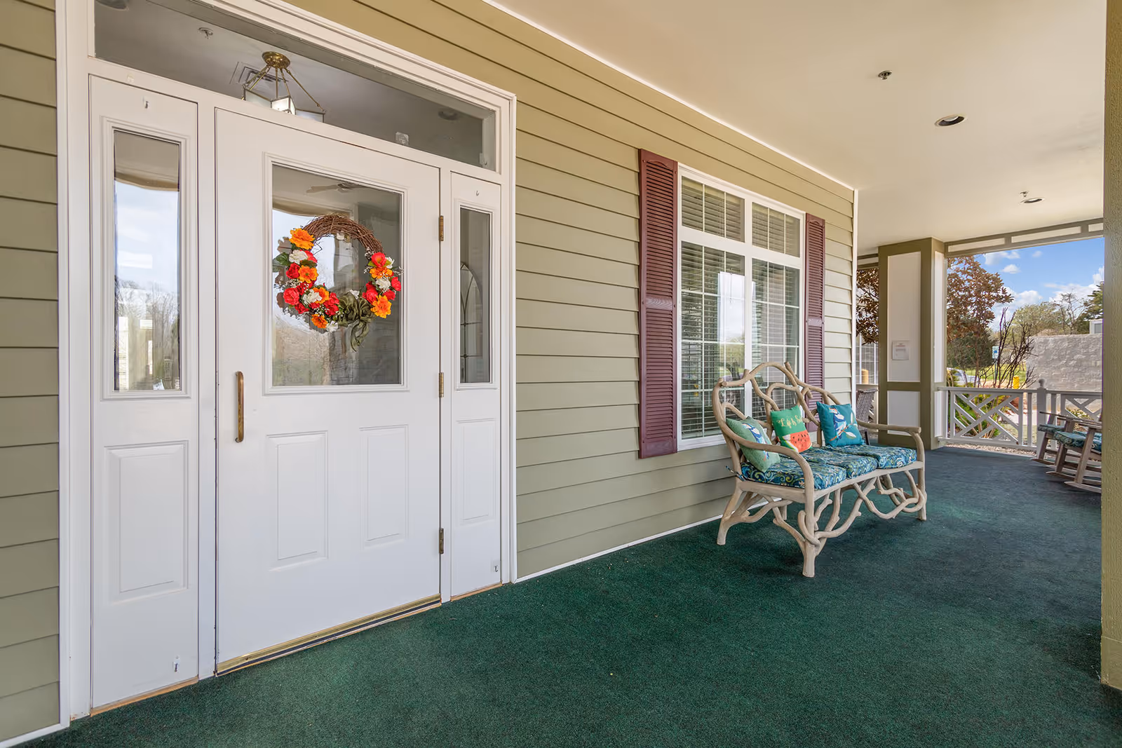 Covered porch area with a white front door decorated with a colorful floral wreath. The porch has green carpeting and a wicker bench with patterned cushions and pillows. There are windows with purple shutters on the wall beside the bench, and rocking chairs are visible further down the porch.