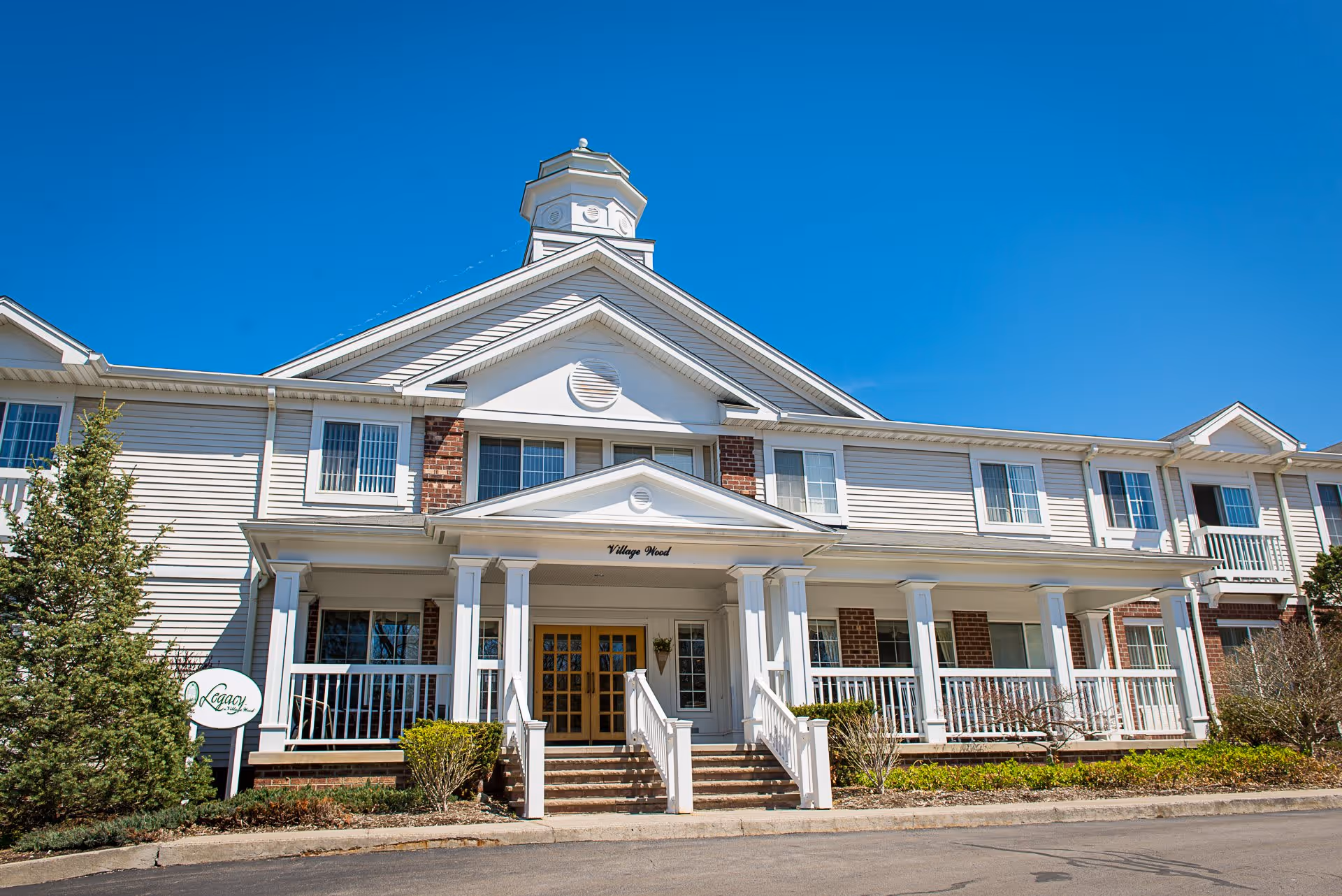 Front exterior view of a two-story senior living facility building named Legacy at Village Wood, featuring white columns, a covered entrance with steps, multiple windows, and a clear blue sky above.