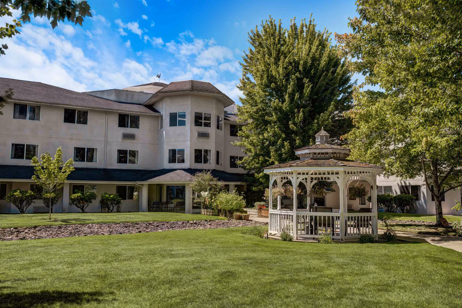 Outdoor view of Holiday Sheldon Oaks facility showing a well-maintained lawn with a white wooden gazebo surrounded by trees and shrubs, and a multi-story building in the background under a partly cloudy blue sky.