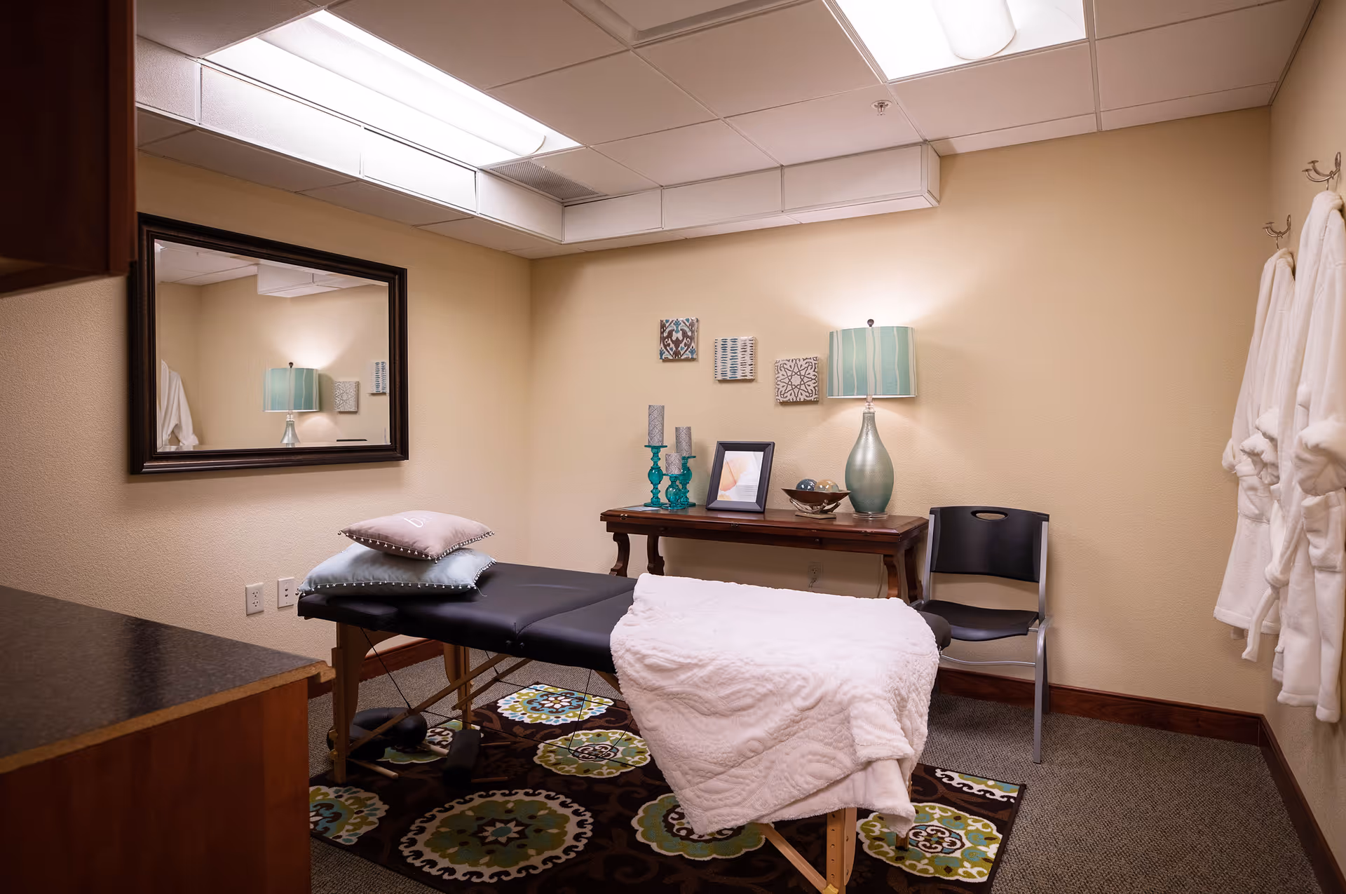 A cozy therapy/massage room with a treatment table draped in a blanket, pillows, a side table with a lamp and decor, a wall mirror, and robes hanging on hooks.