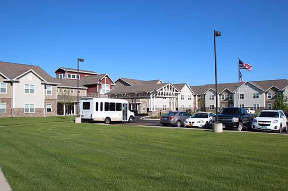 Exterior view of Kennybrook Village senior living facility showing a large green lawn, a parking area with several cars and a shuttle bus, and a multi-story building with beige and red siding under a clear blue sky.