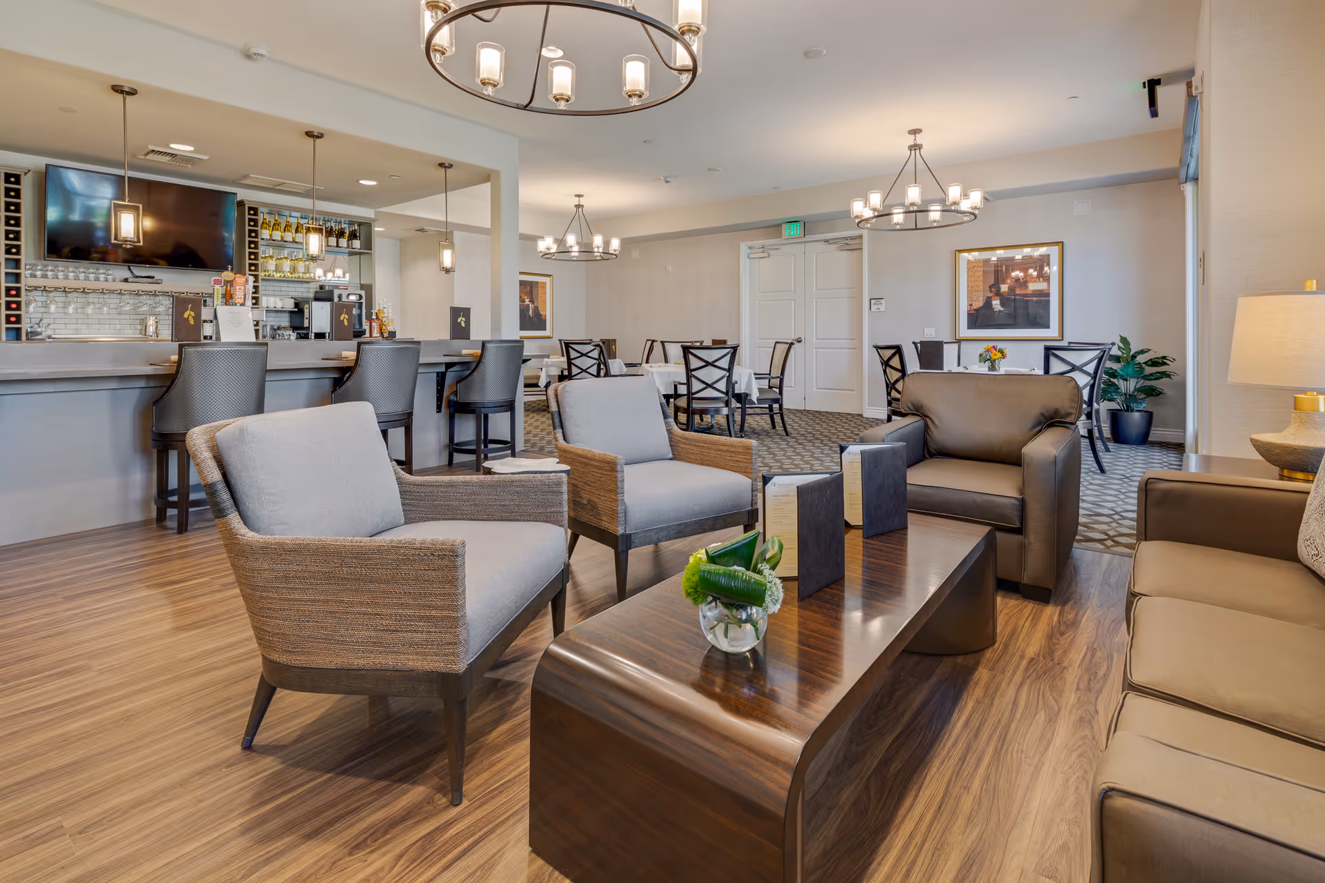 A bright communal lounge with armchairs, a coffee table, bar seating and dining tables in a senior living facility.
