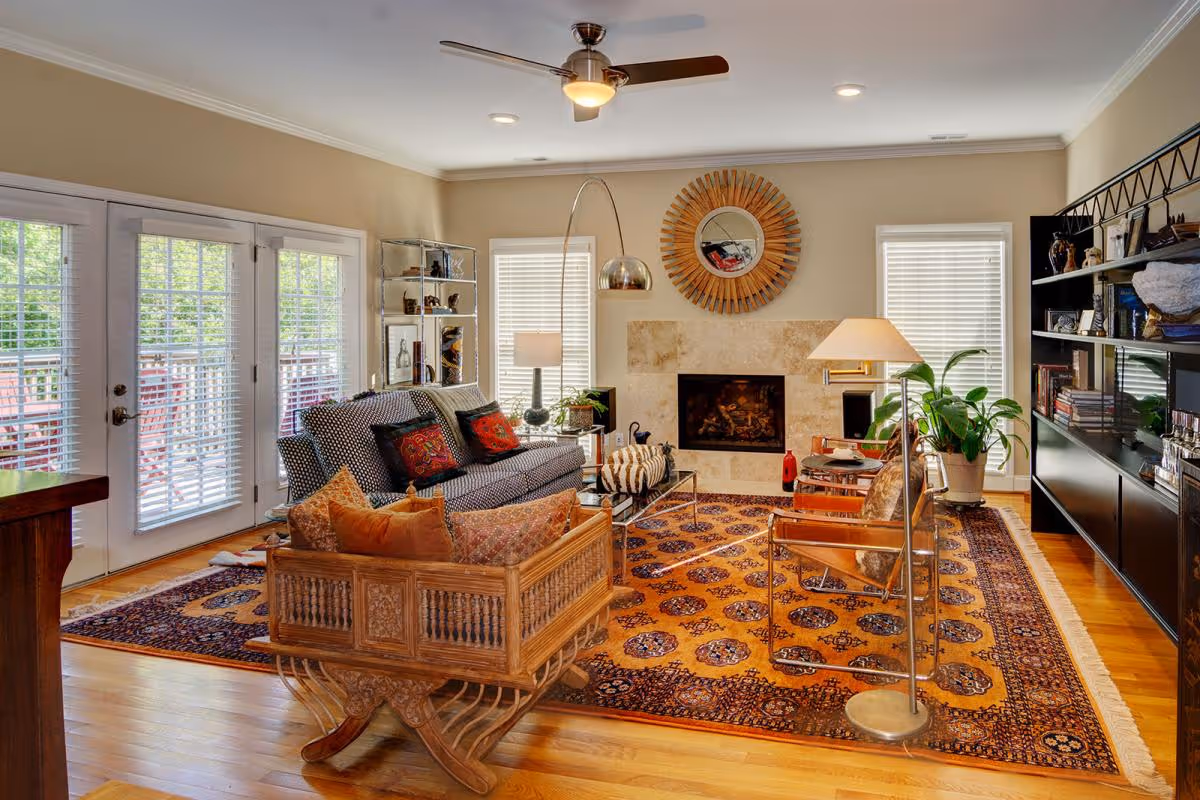 A cozy living room with a patterned orange and blue rug, a wooden carved sofa with orange and patterned cushions, a gray sofa with colorful pillows, two modern chairs with metallic frames, a glass coffee table, a floor lamp, a fireplace with a decorative round mirror above it, large windows with white blinds, a ceiling fan, and a black bookshelf filled with books and decor items.