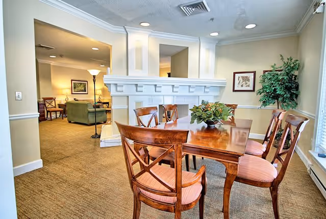 A well-lit interior room featuring a wooden dining table with six chairs upholstered in pink fabric. The room has beige walls, a large window with blinds, a green potted plant in the corner, and a white decorative fireplace with a mirror above it. In the background, there is a glimpse of a living area with green upholstered furniture and a floor lamp.