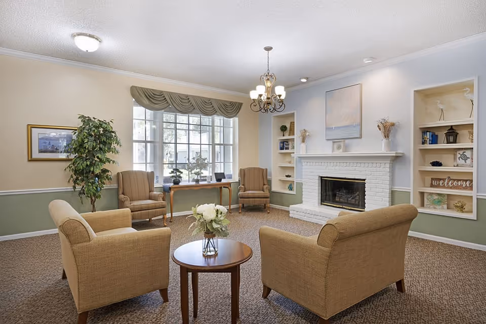 Cozy senior living room with armchairs arranged around a small table facing a white brick fireplace and a large window.
