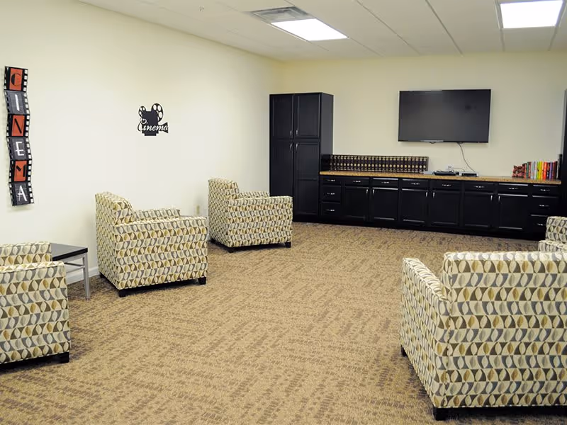 A lounge area with four patterned armchairs arranged around a carpeted floor. On the far wall, there is a large flat-screen TV mounted above a long black cabinet with multiple drawers and cupboards. The walls are light-colored with decorative cinema-themed wall art. The ceiling has recessed lighting.
