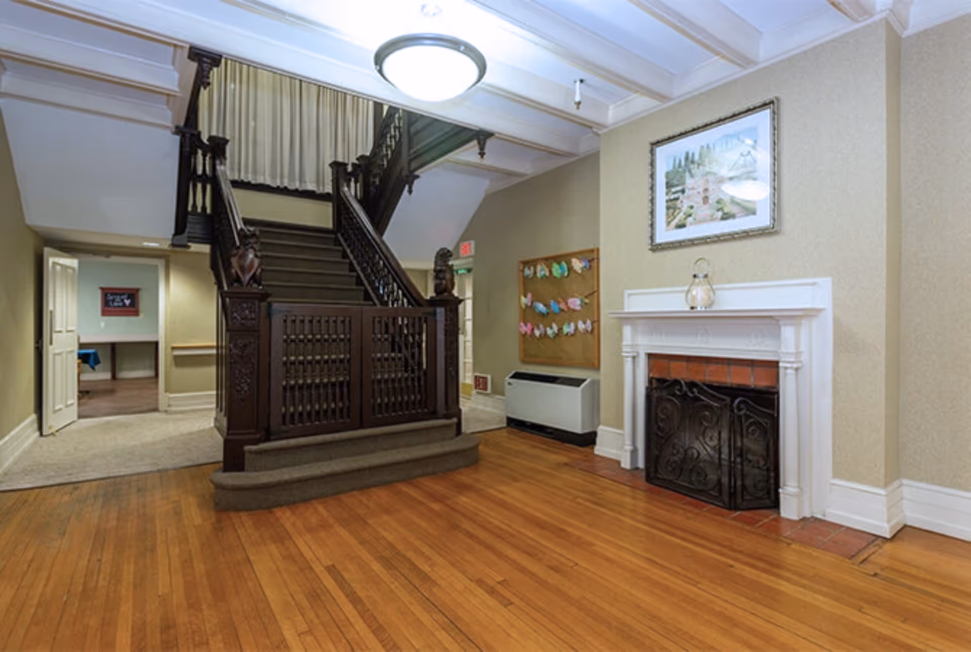 Interior view of a senior living facility featuring a wooden staircase with ornate railings, a white fireplace with a decorative screen, a framed picture above the fireplace, a bulletin board with colorful paper cutouts, and hardwood flooring.