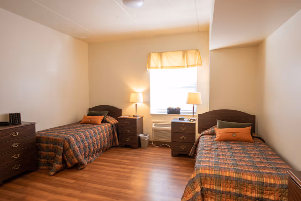 A simple bedroom with two single beds, each with a plaid bedspread in shades of orange and brown, and decorative pillows. Between the beds are two wooden nightstands with lamps. There is a window with a yellow valance letting in natural light. The floor is wood, and the walls are plain and light-colored.