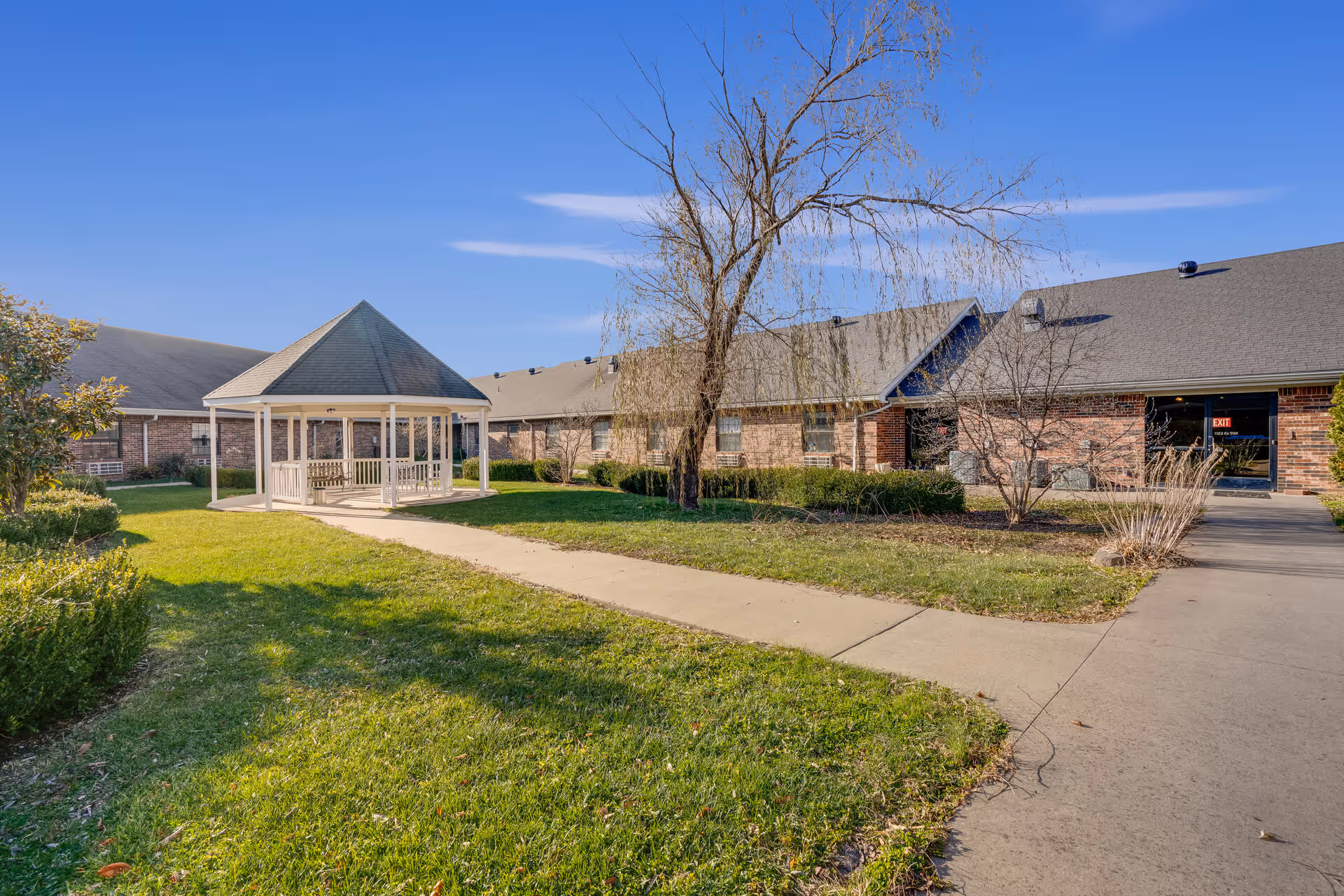 Courtyard of a senior living facility featuring a white gazebo, paved walkways, green lawn, and a brick building in the background.