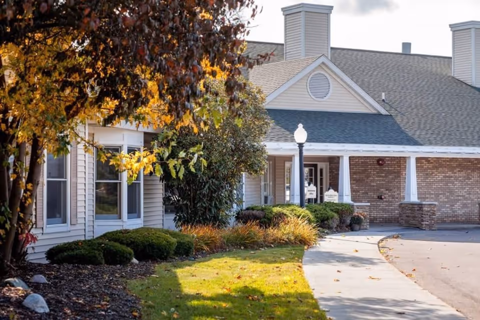 Exterior view of a senior living facility building with beige siding and brick accents, surrounded by landscaped bushes and trees with autumn foliage. A curved sidewalk leads to the entrance under a covered porch with white columns and a streetlamp nearby.