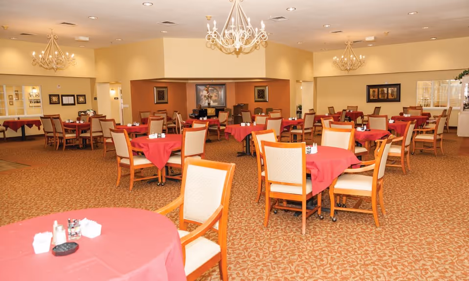 A spacious dining room with multiple round tables covered with red tablecloths and surrounded by wooden chairs with beige cushions. The room has beige walls, patterned carpet, and decorative chandeliers hanging from the ceiling. There are framed artworks on the walls and windows looking into adjacent rooms.