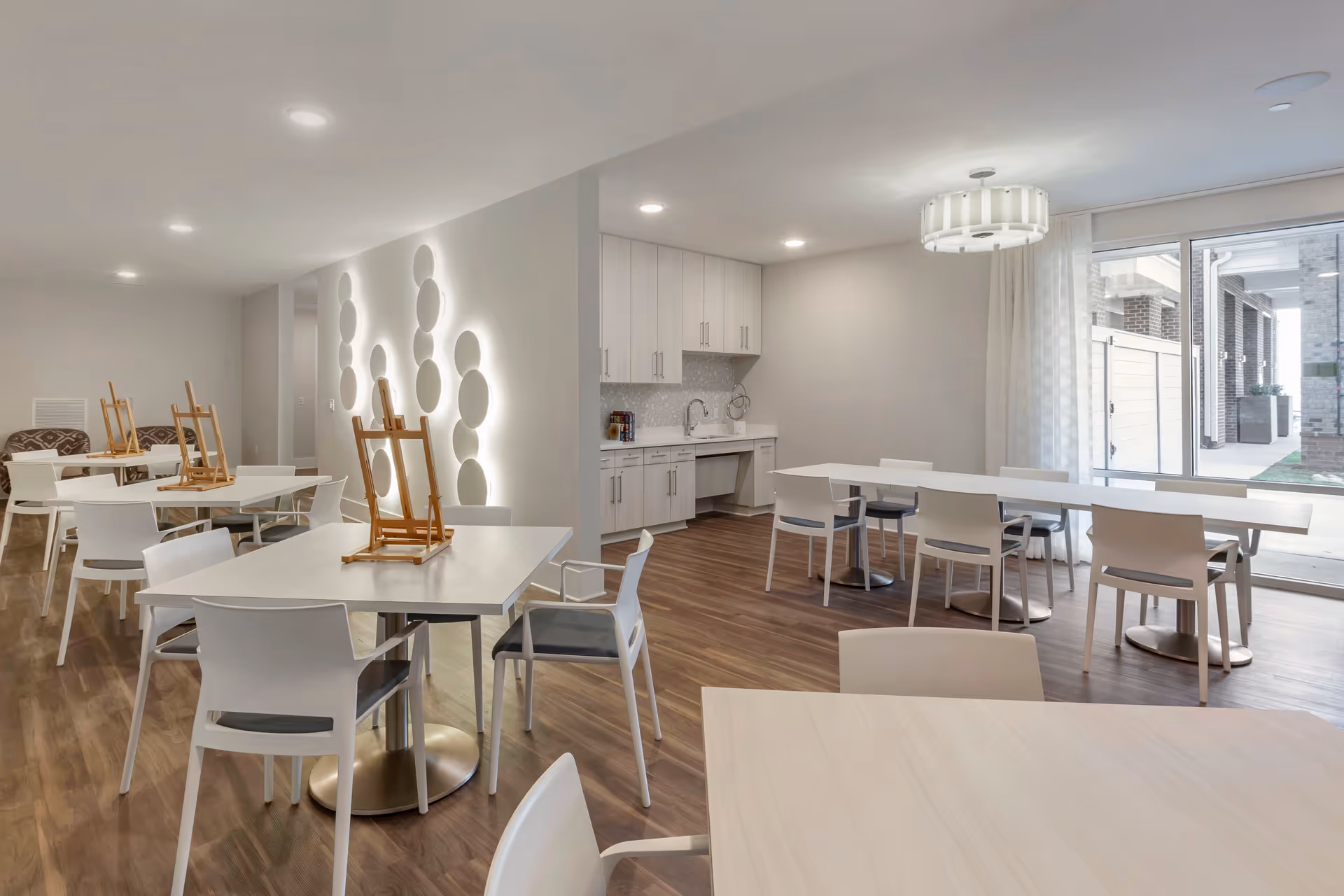 A bright, modern activity room with several white tables and chairs arranged on a wood floor. Some tables have wooden easels on them. The room features a small kitchenette with white cabinets and a sink, a decorative wall with circular light fixtures, and large sliding glass doors leading to an outdoor area.