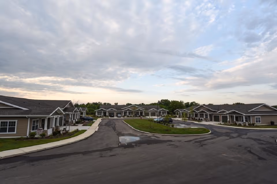 View of a senior living community with single-story residential buildings arranged around a paved road and green landscaped areas under a partly cloudy sky.