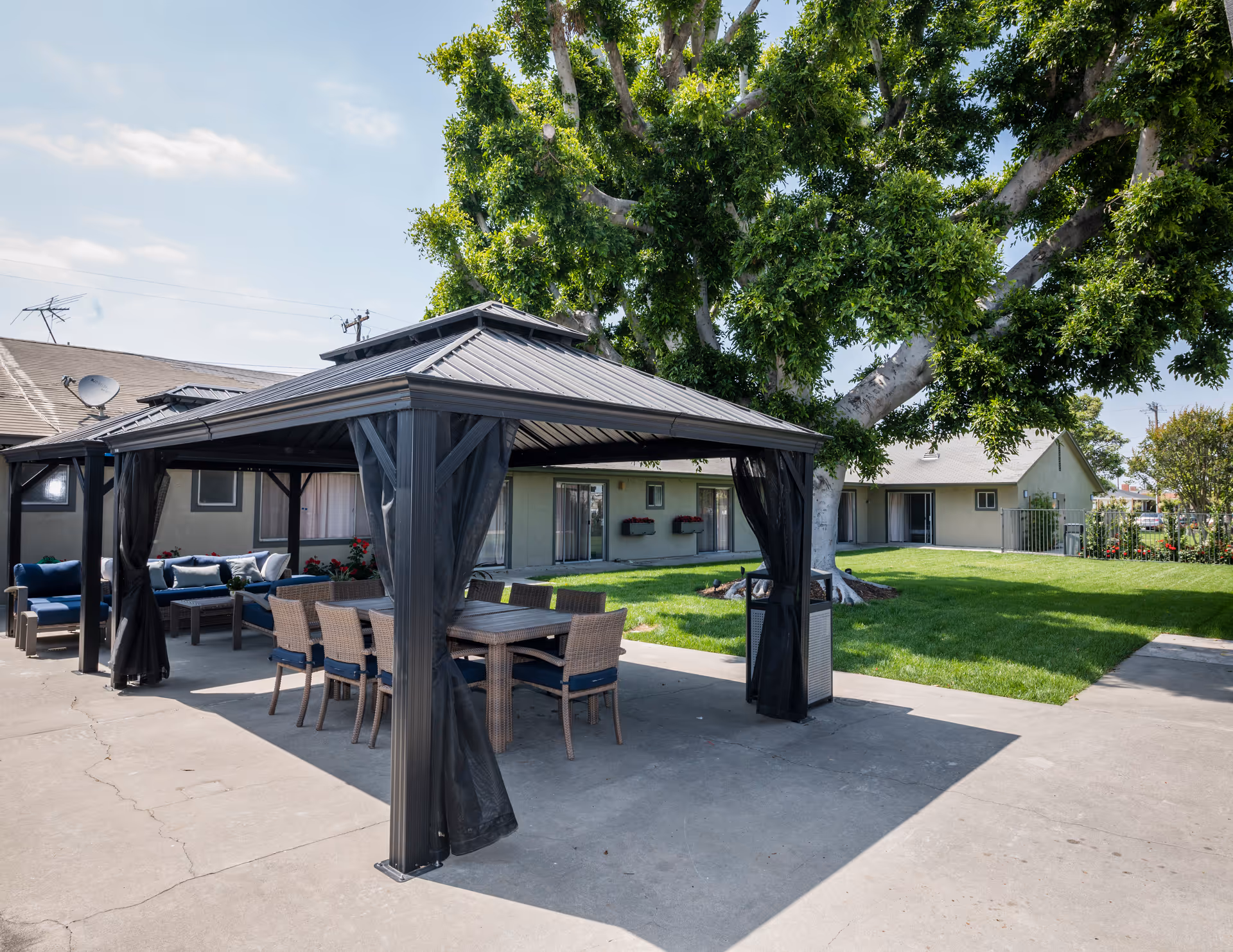 Outdoor seating area at Sun Mar Nursing Center featuring a covered gazebo with a dining table and chairs, surrounded by green grass, large trees, and single-story buildings in the background under a partly cloudy sky.