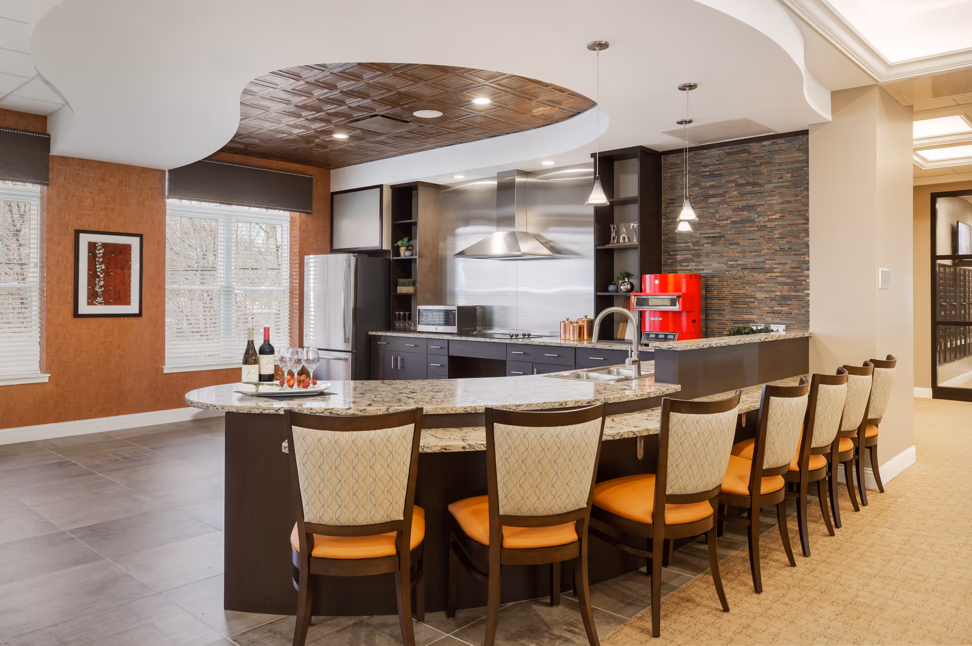 A modern kitchen area in a senior living facility with a long granite countertop island surrounded by eight chairs with orange cushions. The kitchen features stainless steel appliances including a refrigerator and a stove with a vent hood. There is a red coffee machine on the counter and decorative plants on shelves. The ceiling above the kitchen has a decorative wooden panel, and the walls have a mix of textured orange and stone tile finishes. Large windows with white blinds allow natural light into the space.