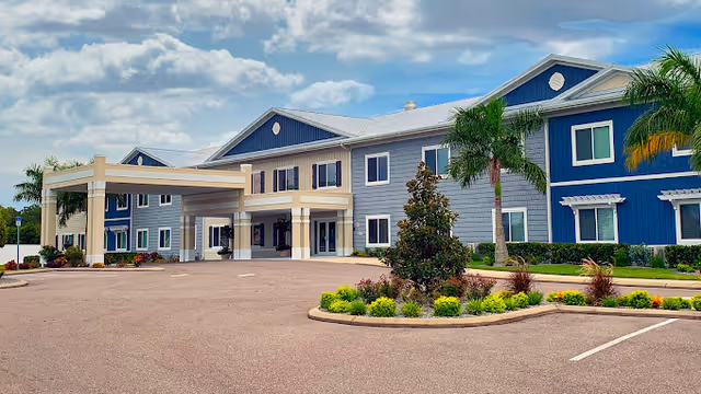 Front exterior of a two-story blue and gray senior living facility with a covered entrance, palm trees, and landscaped circular driveway.