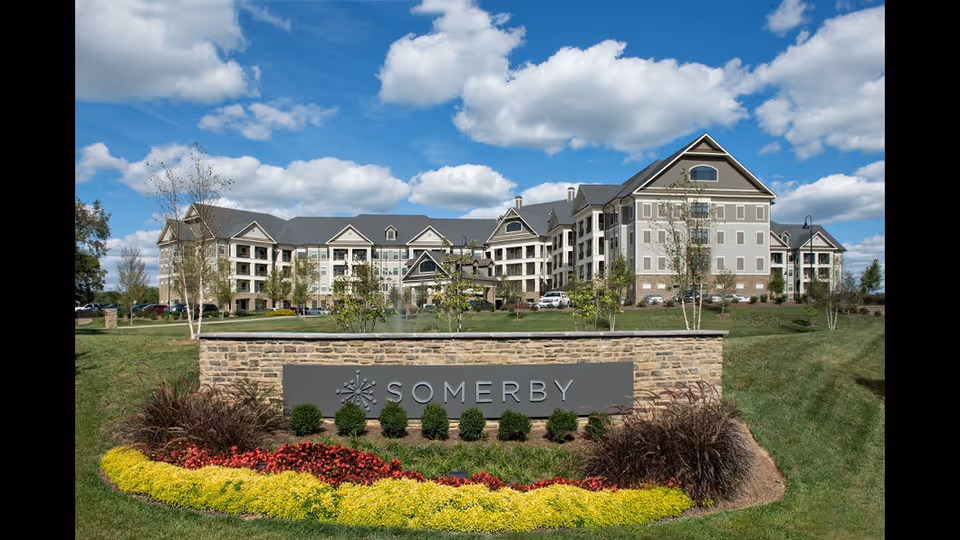 Exterior view of the Somerby Franklin senior living facility building under a partly cloudy blue sky, with a landscaped stone sign in the foreground surrounded by colorful flowers and greenery.