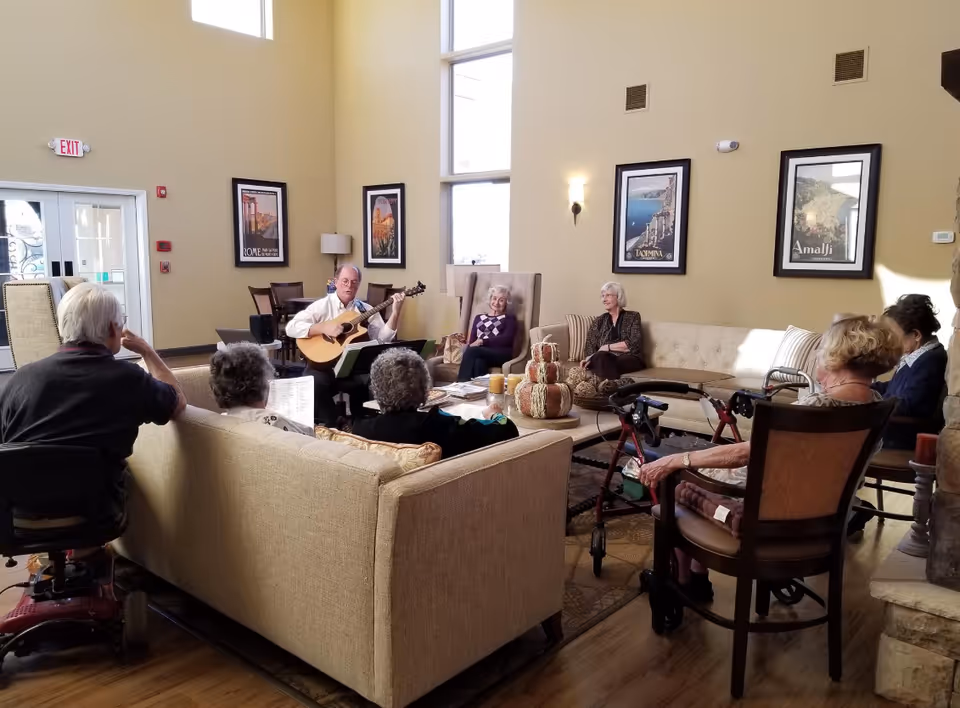 A group of elderly people seated in a spacious, well-lit living room with high ceilings and large windows. One man is playing a guitar while others listen attentively, sitting on sofas and chairs arranged in a circle. The room has framed pictures on the walls and a decorative centerpiece on the coffee table.