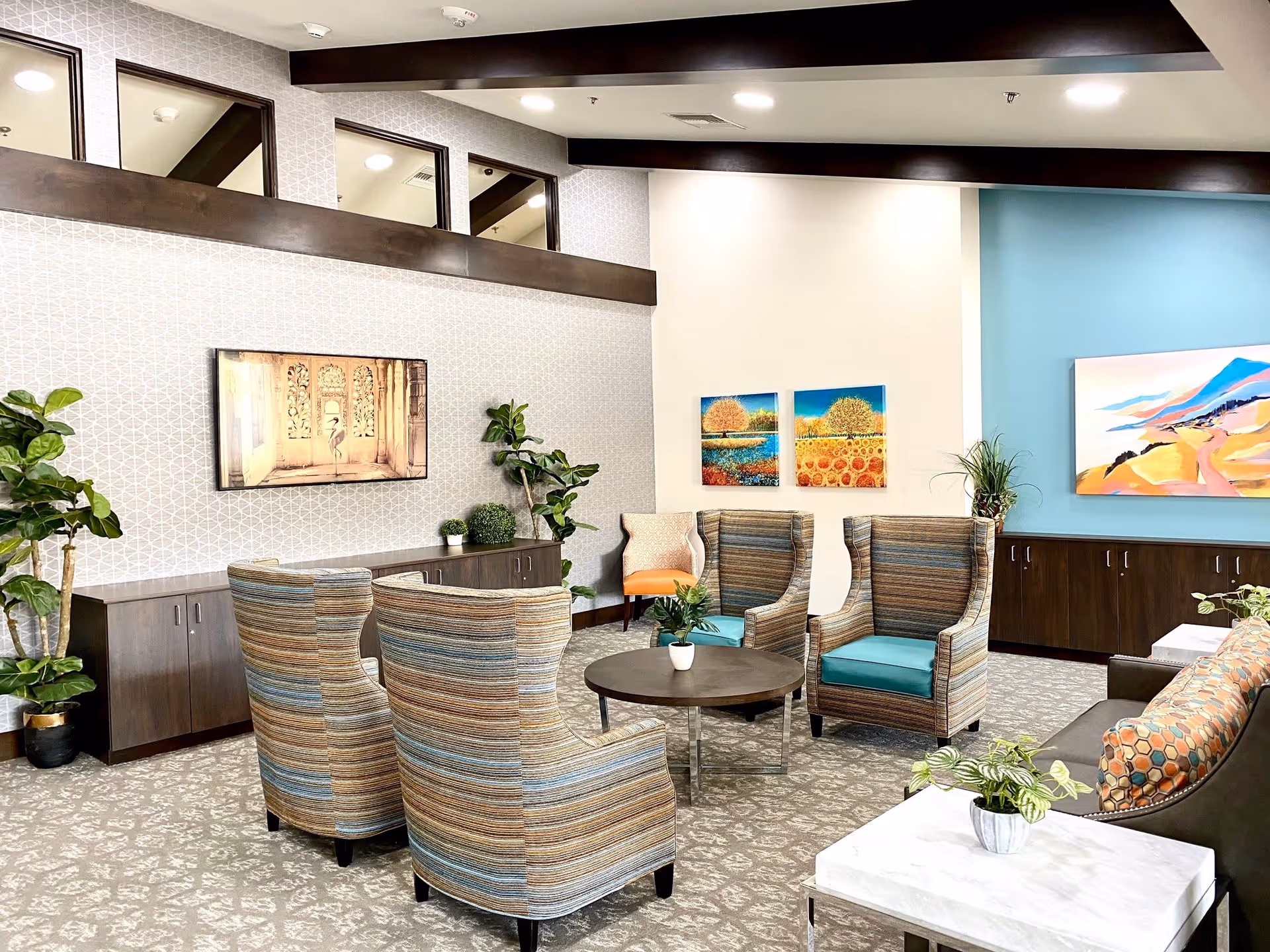 A bright and modern living room area with patterned carpet, four striped armchairs with teal cushions arranged around a round coffee table, a dark brown sofa with a colorful pillow, and several potted plants. The walls feature geometric wallpaper, three colorful paintings, and dark wood cabinetry along the back wall. The ceiling has dark wood beams and recessed lighting.