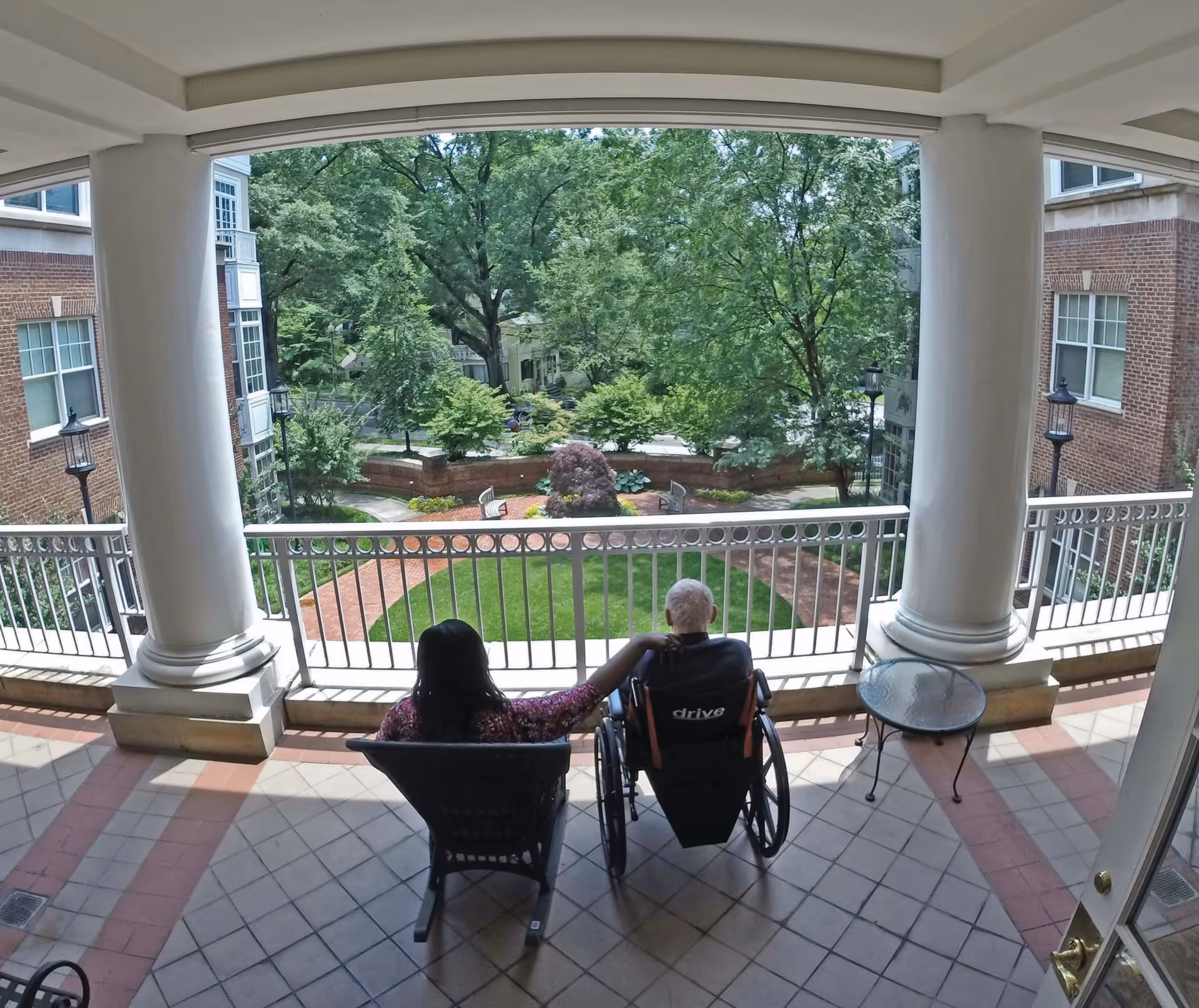 Two people sitting on a covered balcony overlooking a landscaped garden with trees, shrubs, and benches. One person is in a wheelchair and the other is seated in a chair with their arm around the person in the wheelchair. The balcony has large white columns and a small round glass table.