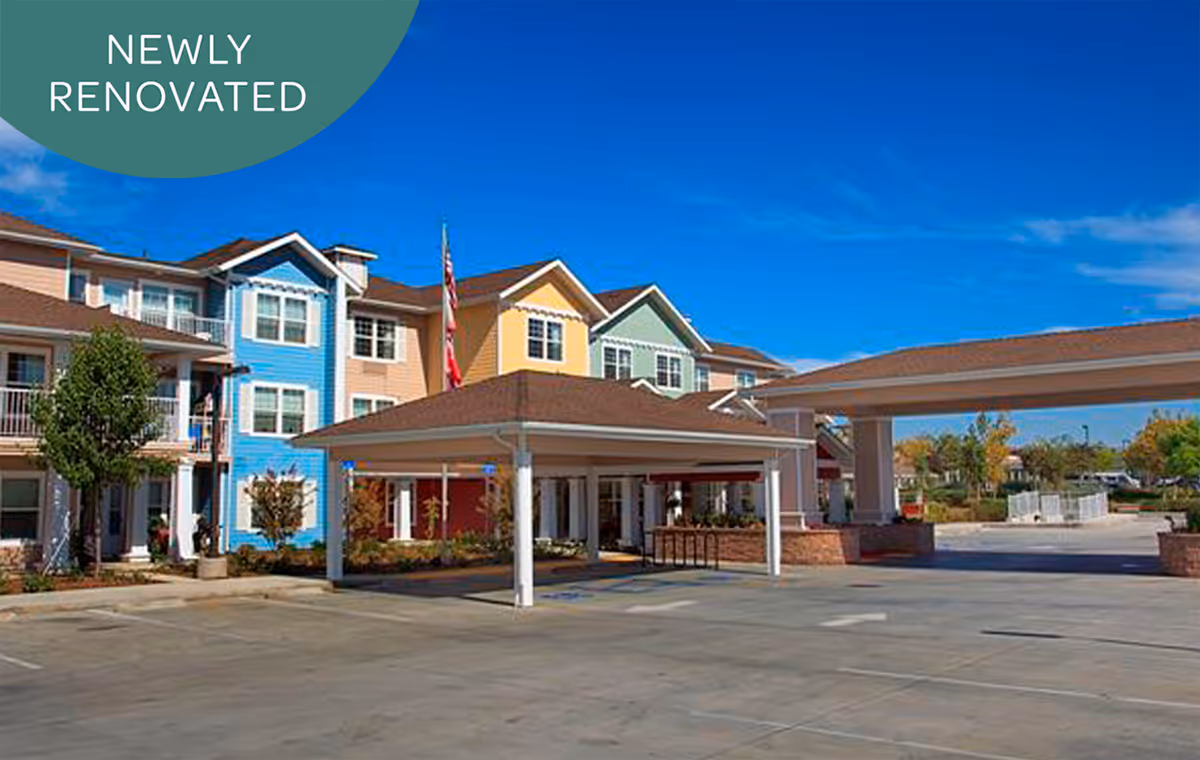 Exterior view of a senior living facility with colorful buildings in blue, yellow, and green. There is a covered drop-off area in front of the entrance, a clear blue sky, and a green banner in the top left corner that says 'NEWLY RENOVATED'.