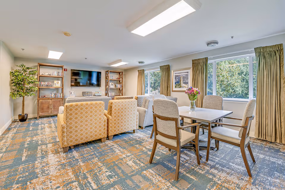 A bright and cozy senior living common area with patterned armchairs arranged facing a wall-mounted flat-screen TV. There are two wooden bookshelves flanking the TV, a potted plant in the corner, and a table with four chairs and a vase of flowers near large windows with beige curtains letting in natural light.