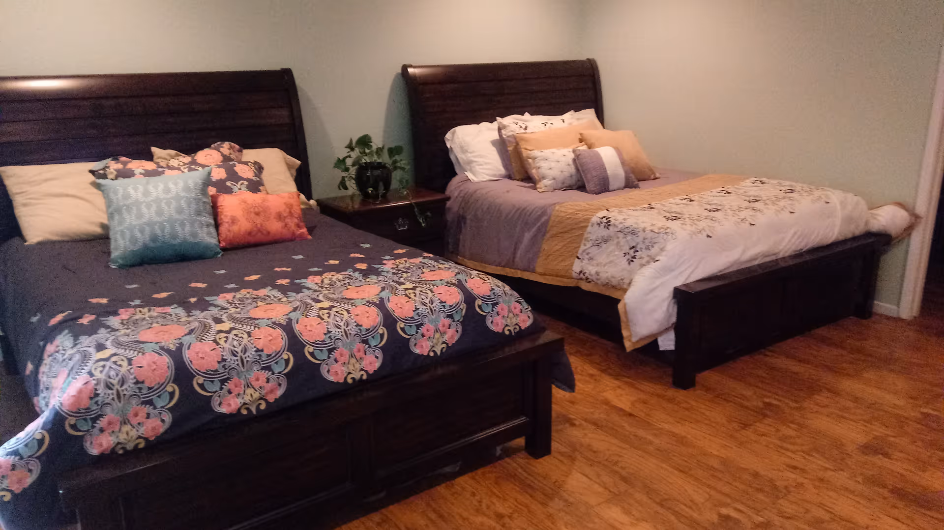 A bedroom with two dark wooden beds, each made with colorful bedding and multiple pillows. There is a small wooden nightstand with a green plant between the beds. The room has wooden flooring and light-colored walls.